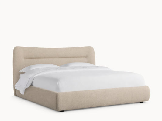 Lille Bed in fawn textured fabric with a curved upholstered headboard featuring horizontal lines, white bedding, on a wood floor.