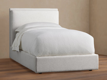 Lennon Twin Bed in light fabric with a high upholstered headboard, white bedding, on a wooden floor in a neutral-colored room.