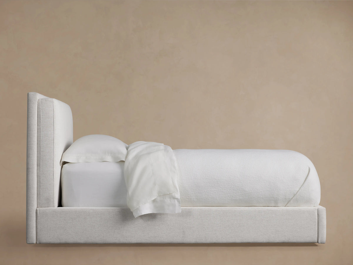 Lennon Twin Bed with light beige upholstery, side profile view. Includes plush headboard, white bedding neatly tucked in.