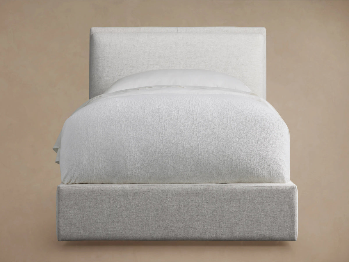 Twin size Lennon bed with a soft beige upholstered headboard and frame, neatly made with white bedding, shown against a light brown wall.