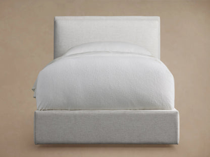 Twin size Lennon bed with a soft beige upholstered headboard and frame, neatly made with white bedding, shown against a light brown wall.
