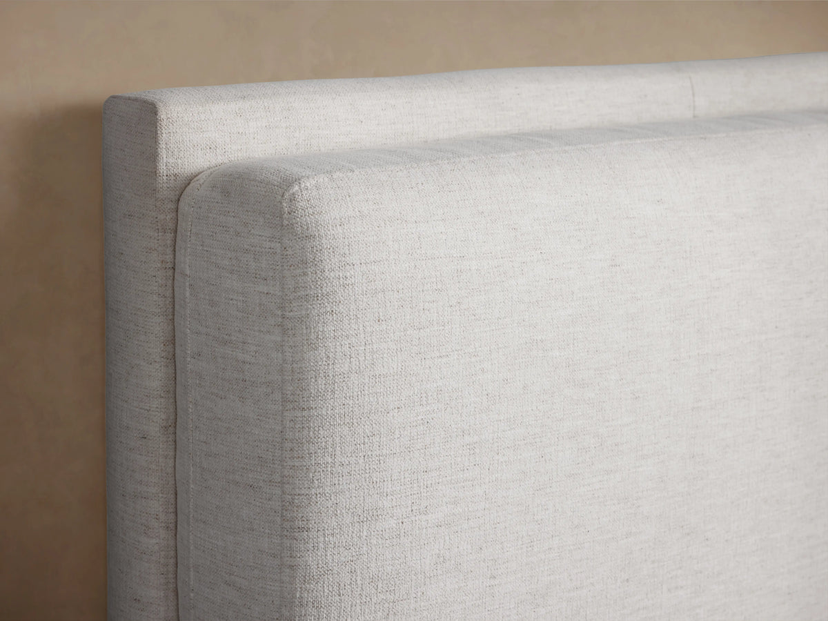 Lennon Twin Bed in light fabric with a high upholstered headboard, white bedding, on a wooden floor in a neutral-colored room.