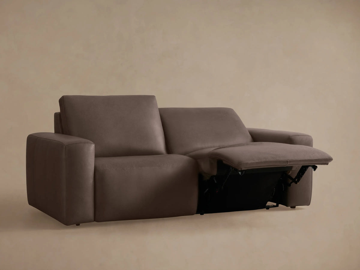 Brennan Leather Two-Piece Motion Sofa in rich brown/taupe. Left seat reclined with footrest extended, against a warm neutral wall.