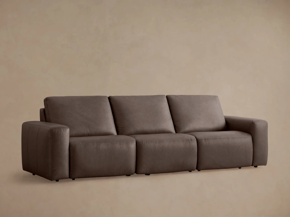 Front view of Brennan Leather Three-Piece Motion Sofa in deep taupe, featuring three connected recliner sections, against a simple tan wall & wood floor.