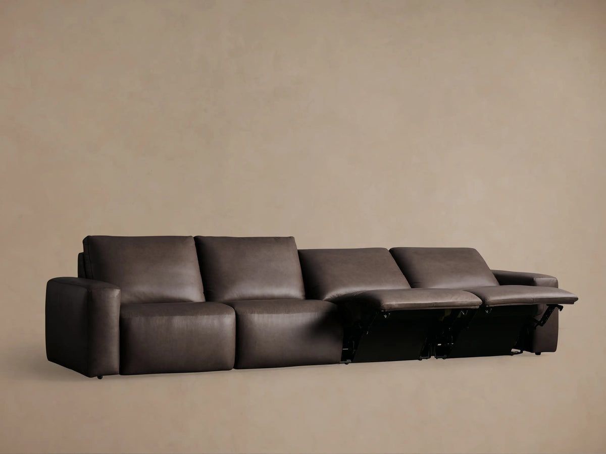 Dark brown Brennan Leather Four-Piece Motion Sectional, one seat reclined with footrest extended, against a light brown wall.