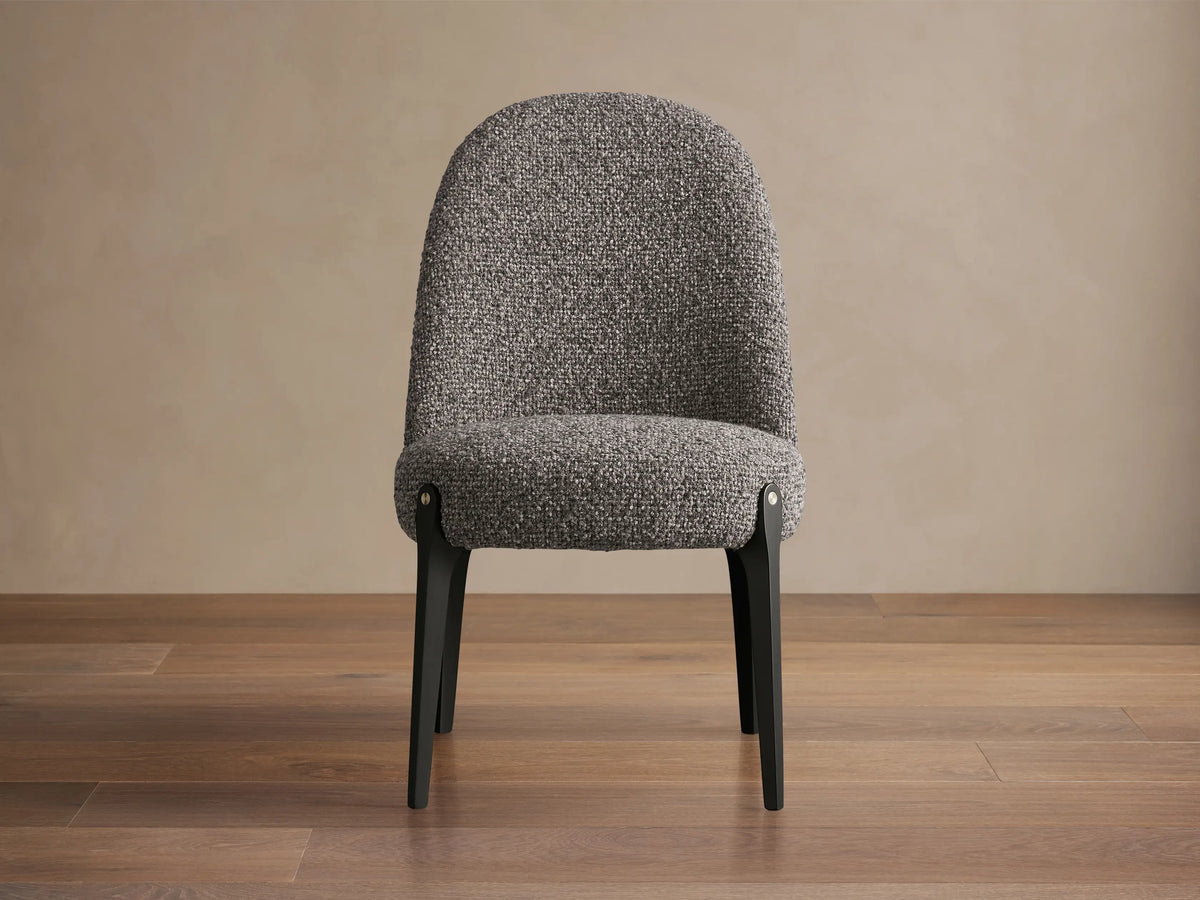 Bottoni Dining Chair, gray textured upholstery, rounded back, dark legs with brass tack detail, on wooden floor.