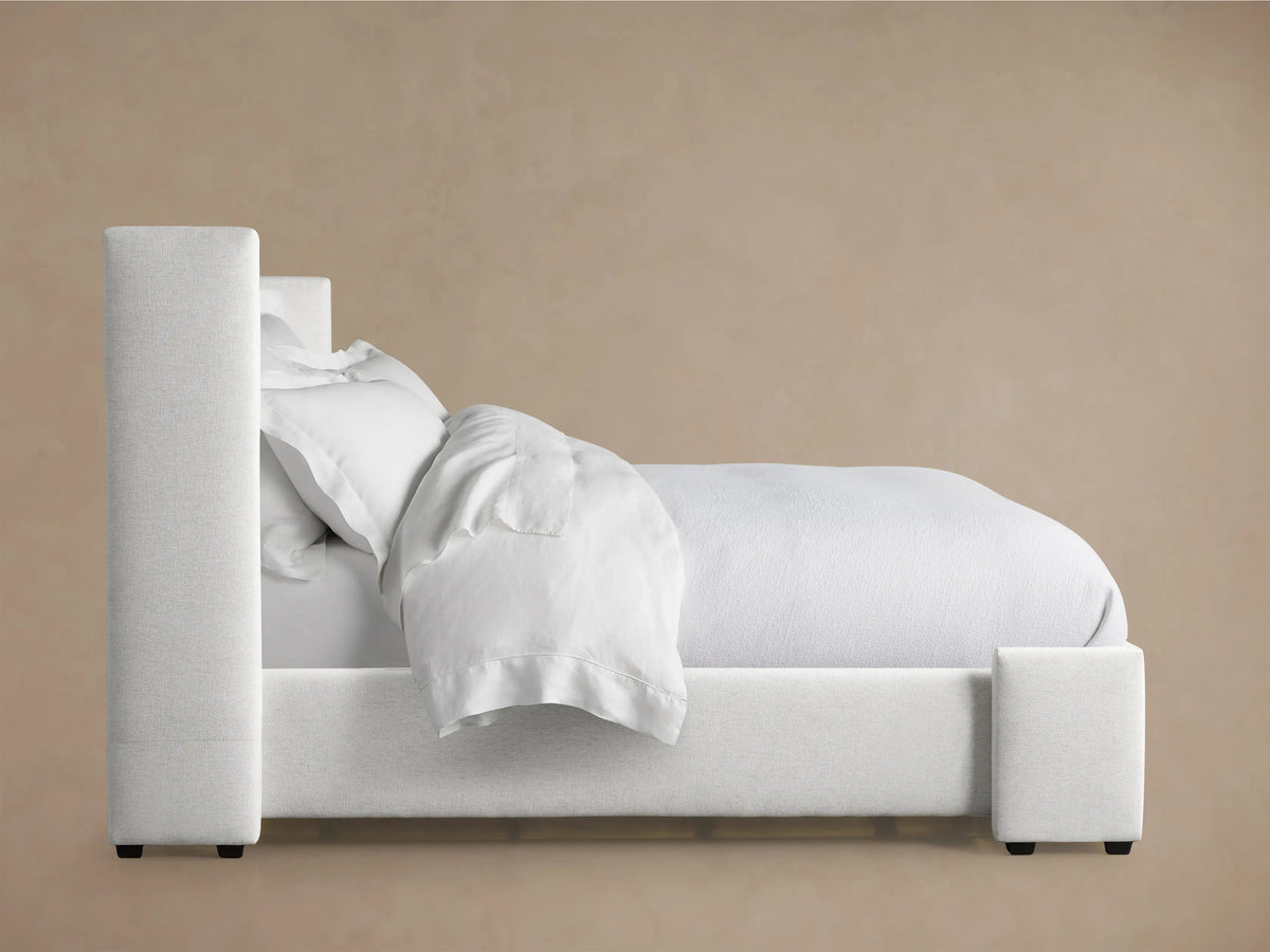 Wyller Box-Tufted Bed headboard. Close-up of the light gray, textured fabric with square tufting against a brown wall.