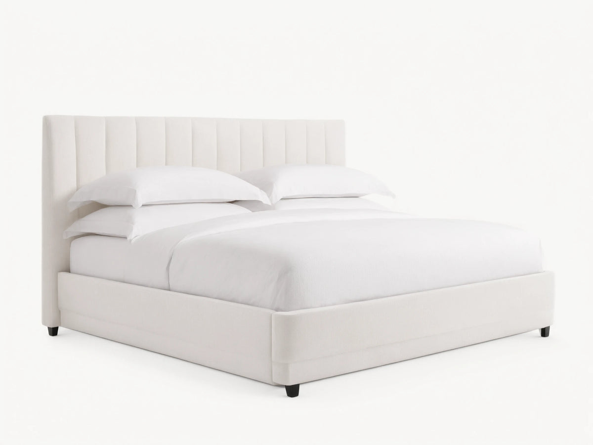 Wyller channel-stitched extended headboard bed in white, with low footboard, white bedding, on wood floor against beige wall.