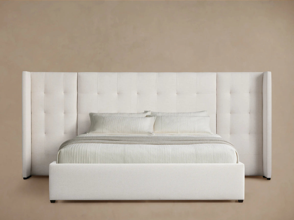 Off-white Wyller bed with box-tufted, extended headboard & low footboard. Light bedding on wooden floor, angled view.