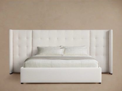 Off-white Wyller bed with box-tufted, extended headboard & low footboard. Light bedding on wooden floor, angled view.