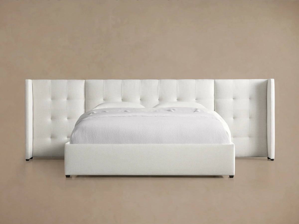 White Wyller Box-Tufted Extended Headboard Bed, upholstered with low footboard & white bedding, angled against a light brown wall.