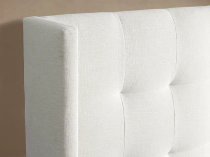 Side view of Wyller Extended Headboard Bed. Light neutral upholstered frame, white bedding, against a light brown wall.