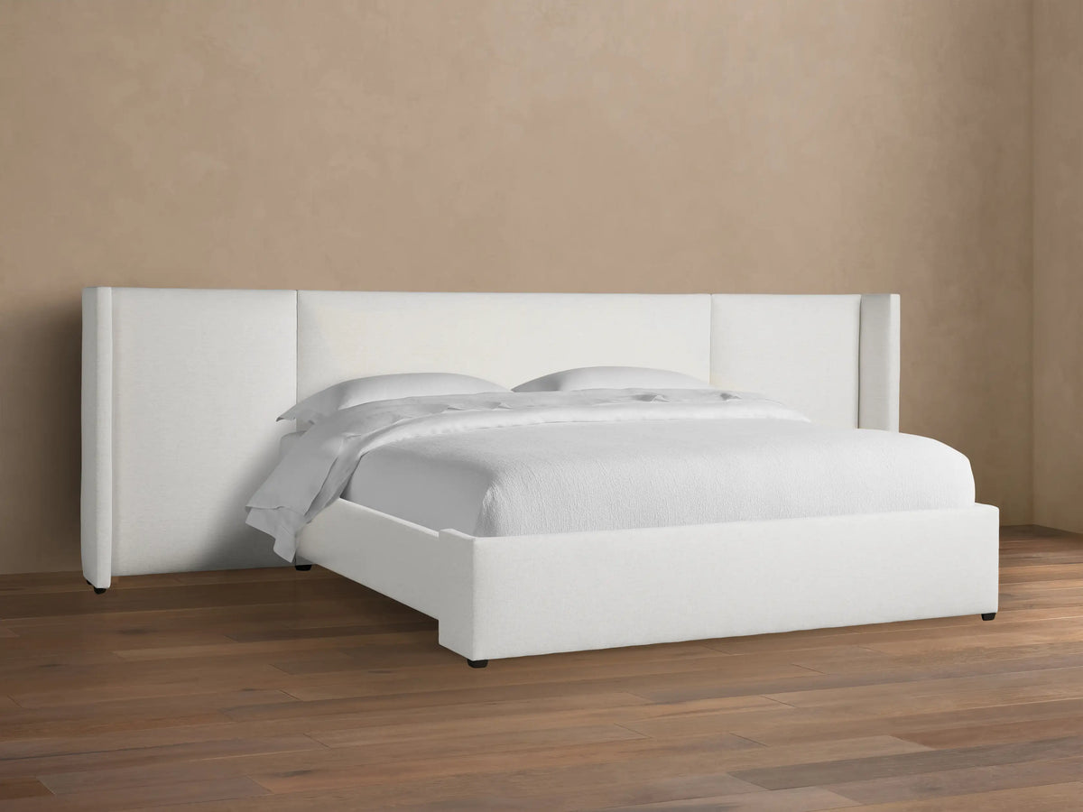 White upholstered Wyller Extended Headboard Bed with low footboard, white bedding, on wood floor against tan wall.