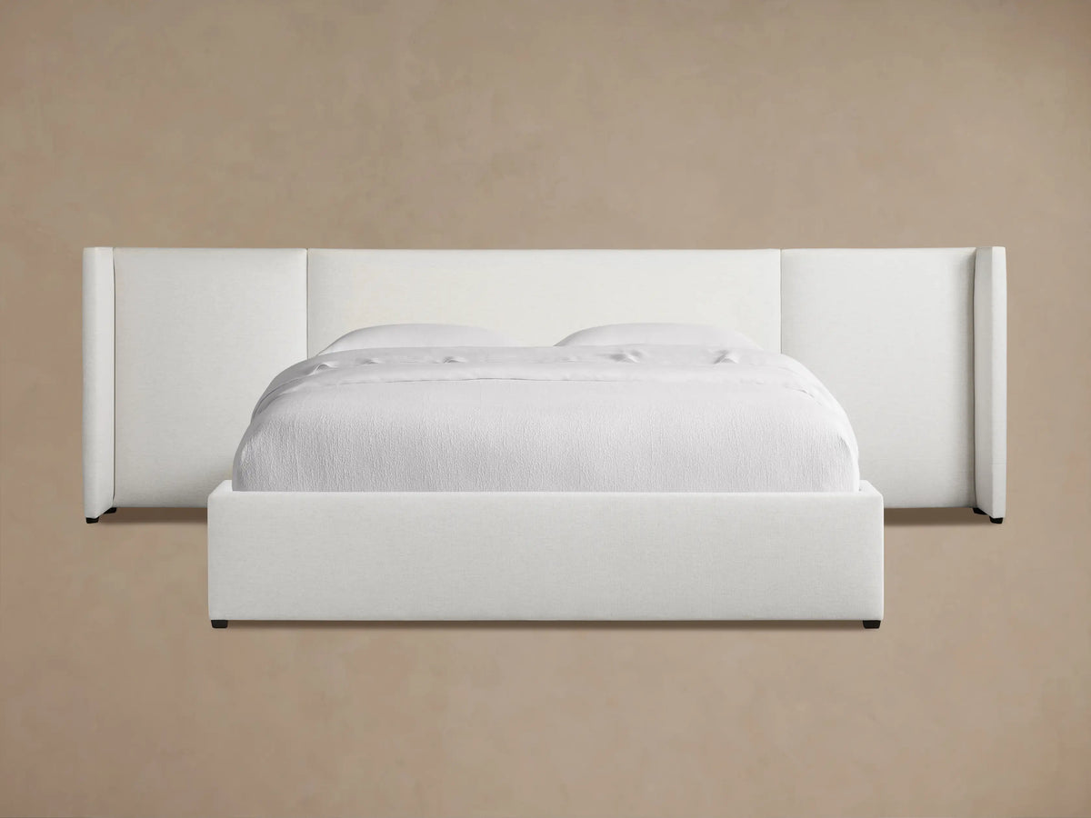 White upholstered Wyller Extended Headboard Bed with low footboard, white bedding, on wood floor against tan wall.