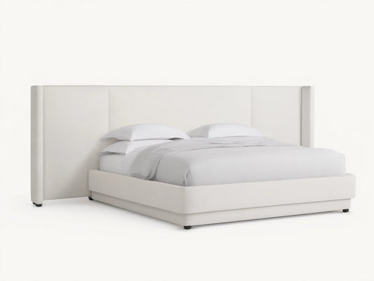 White upholstered Wyller Extended Headboard Bed with low footboard, white bedding, on wood floor against tan wall.