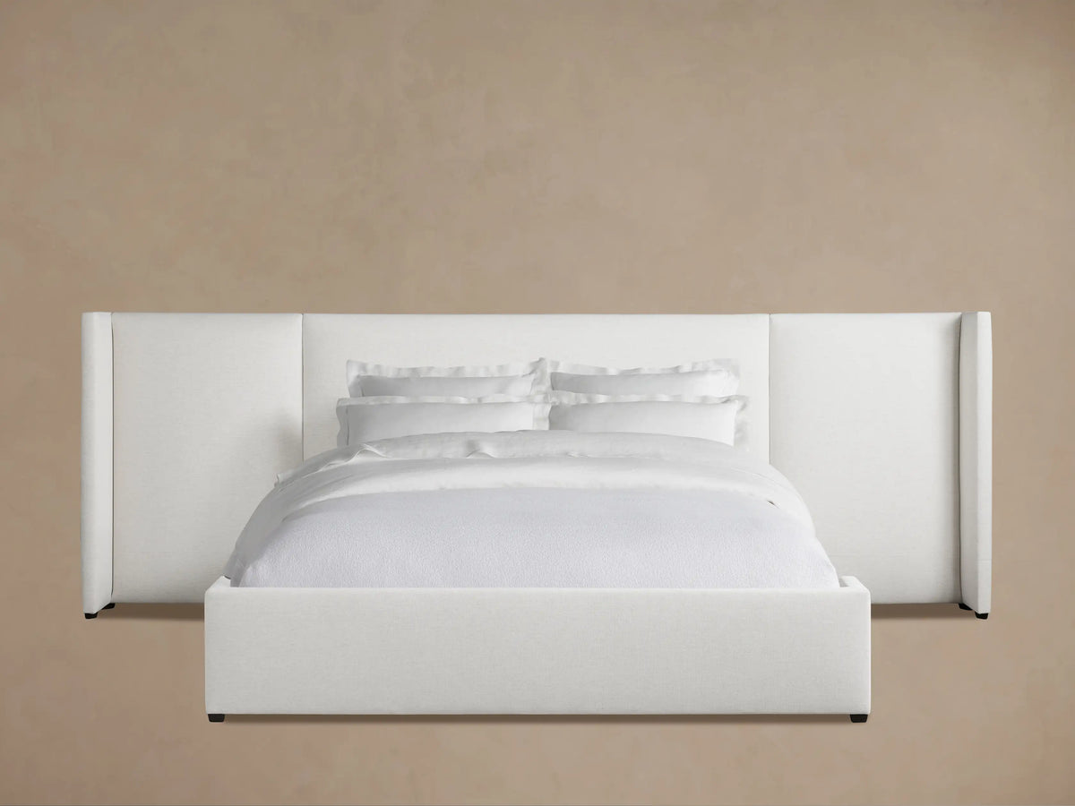 White upholstered Wyller Extended Headboard Bed with low footboard, made up with white bedding, against a tan wall & wood floor.
