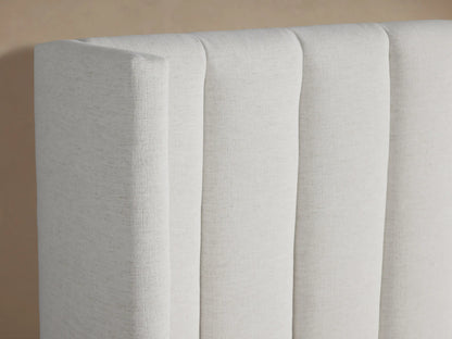 Wyller Channel-Stitched Bed. White upholstered bed with tall, vertically stitched headboard, low footboard & white bedding. On beige wall.