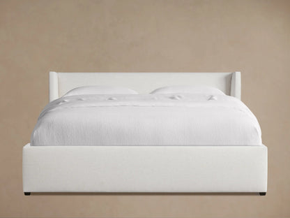 Angled view of a white upholstered Wyller Bed with a wingback headboard and white bedding on a hardwood floor.