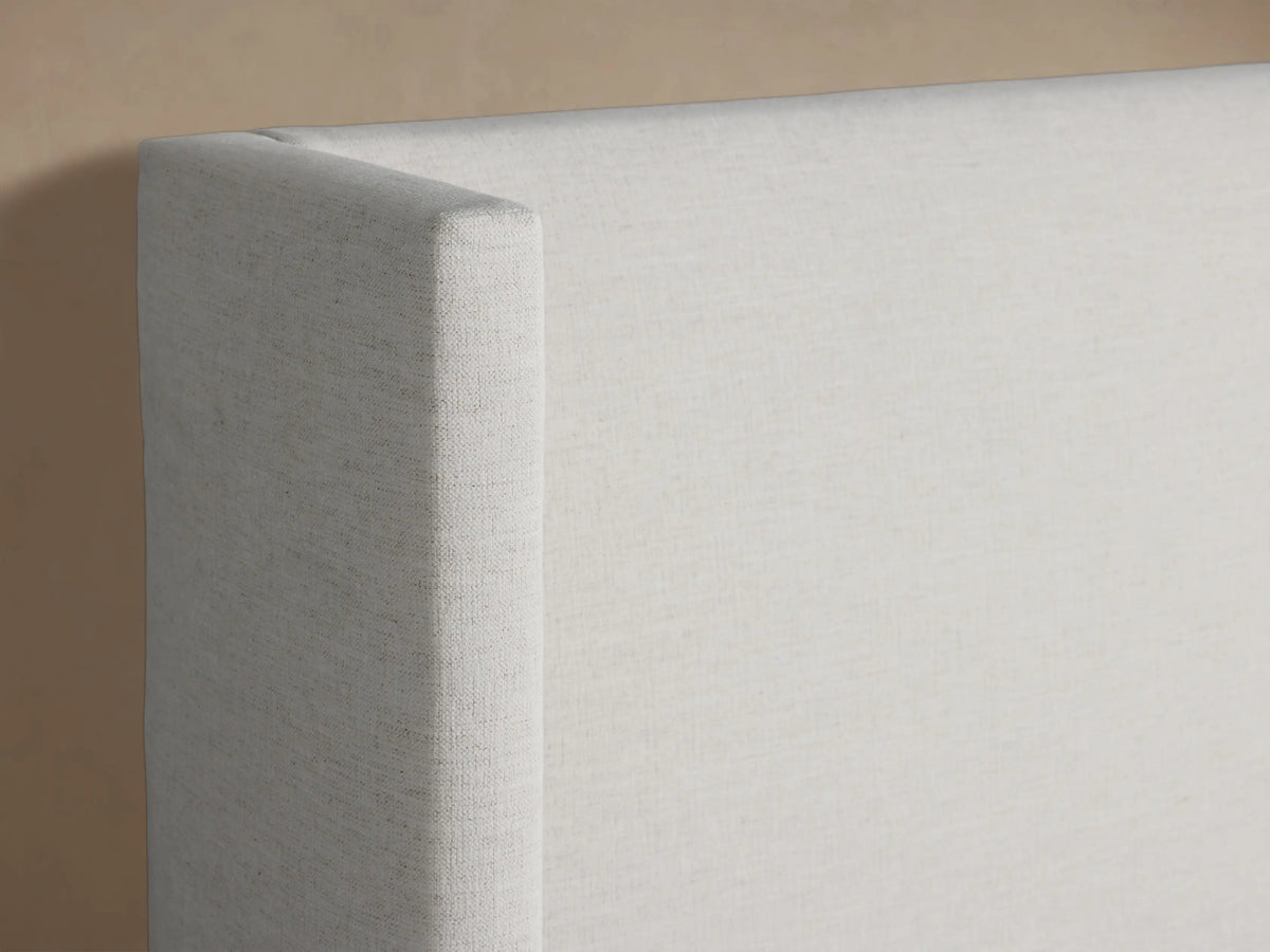 Front view of a Wyller Bed, upholstered in light cream fabric with a tall winged headboard and white bedding, against a plain beige wall.