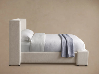 Wyller Bed, upholstered in oatmeal textured fabric, with white bedding and a light blue throw. Side view.
