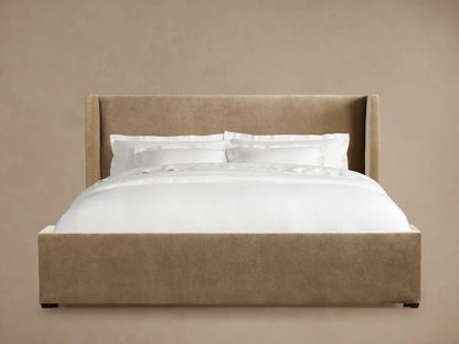 Wyller Bed with tan upholstered frame and winged headboard, dressed with white pillows and duvet, against a tan wall.
