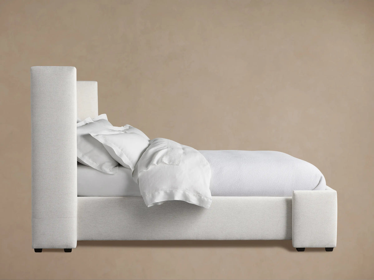 Wyller Bed upholstered in light off-white fabric, side view. Features tall rectangular headboard & footboard with white bedding.