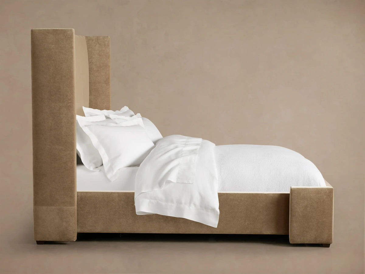 Wyller Bed: Close-up of an upholstered headboard corner in soft, light brown, velvety fabric against a warm beige wall.