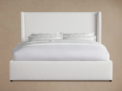 White upholstered Wyller Bed with a high winged headboard and matching footboard, neatly made with white bedding, on wood floor.