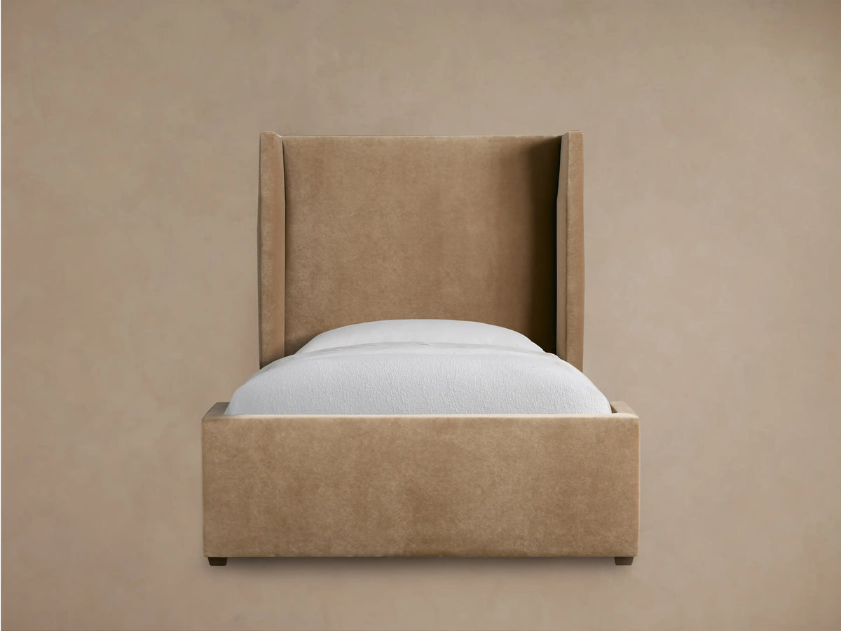 Wyller Twin Bed, upholstered in tan fabric, tall wingback headboard, white bedding, on a wooden floor against a beige wall.