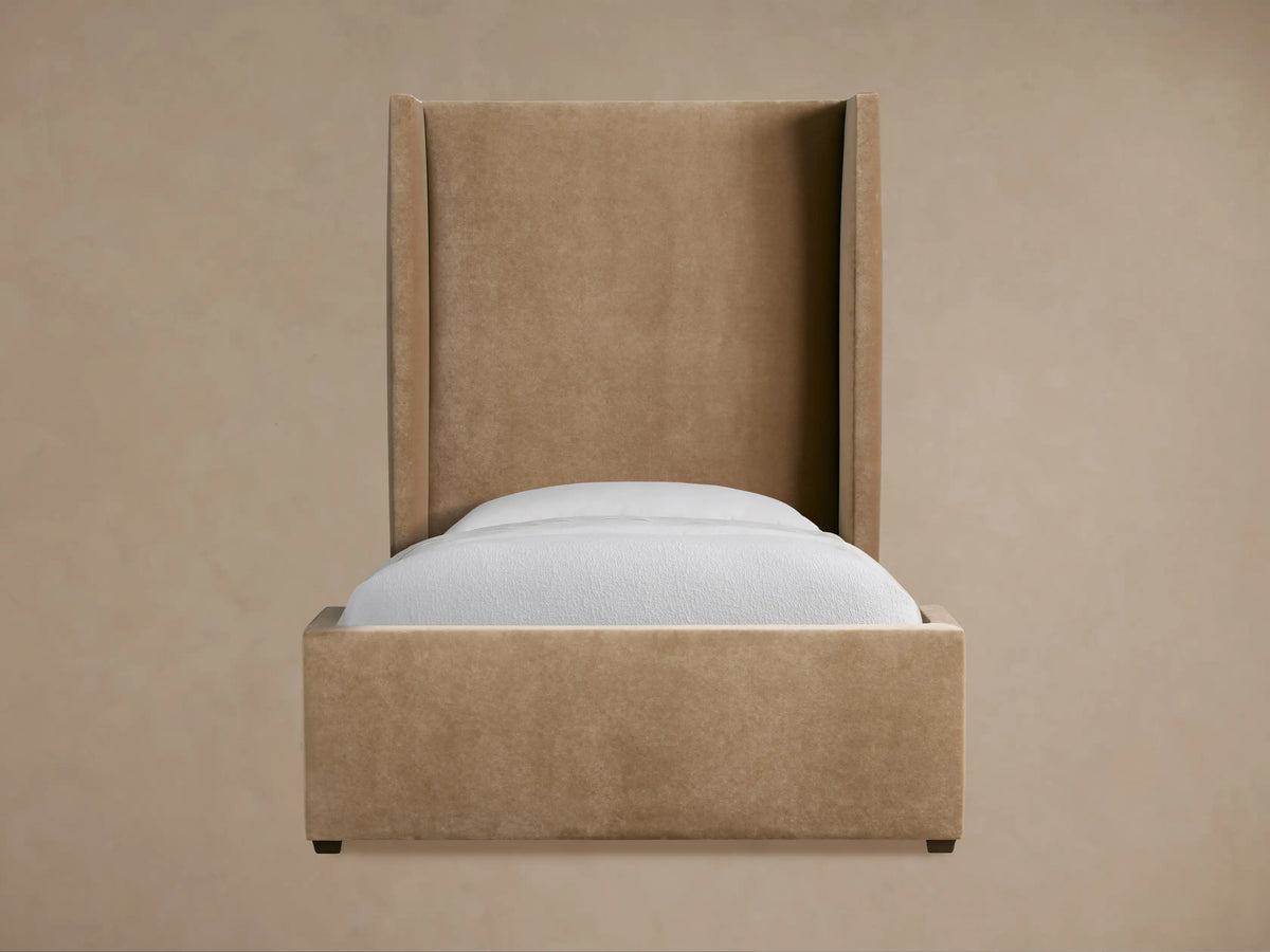 Tan upholstered Wyller Twin Bed with tall winged headboard and white bedding, viewed from an angle in a neutral room.