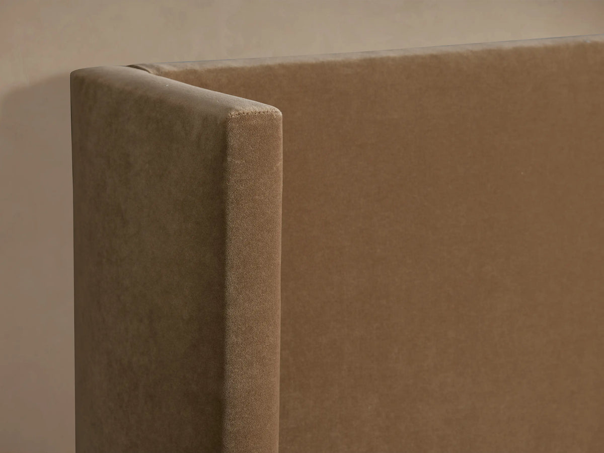 Wyller Twin Bed. Tan upholstered frame with a tall, winged headboard and white bedding, against a beige wall.