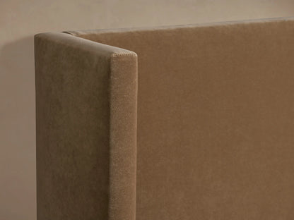 Wyller Twin Bed. Tan upholstered frame with a tall, winged headboard and white bedding, against a beige wall.