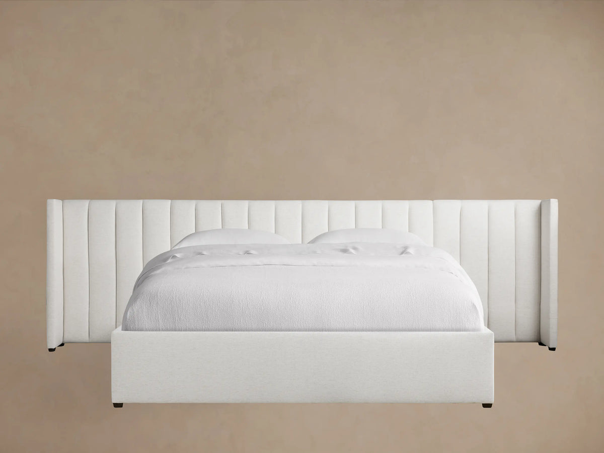 White Wyller Channel-Stitched Extended Headboard Bed with low footboard, white bedding, on a dark wood floor against a tan wall.