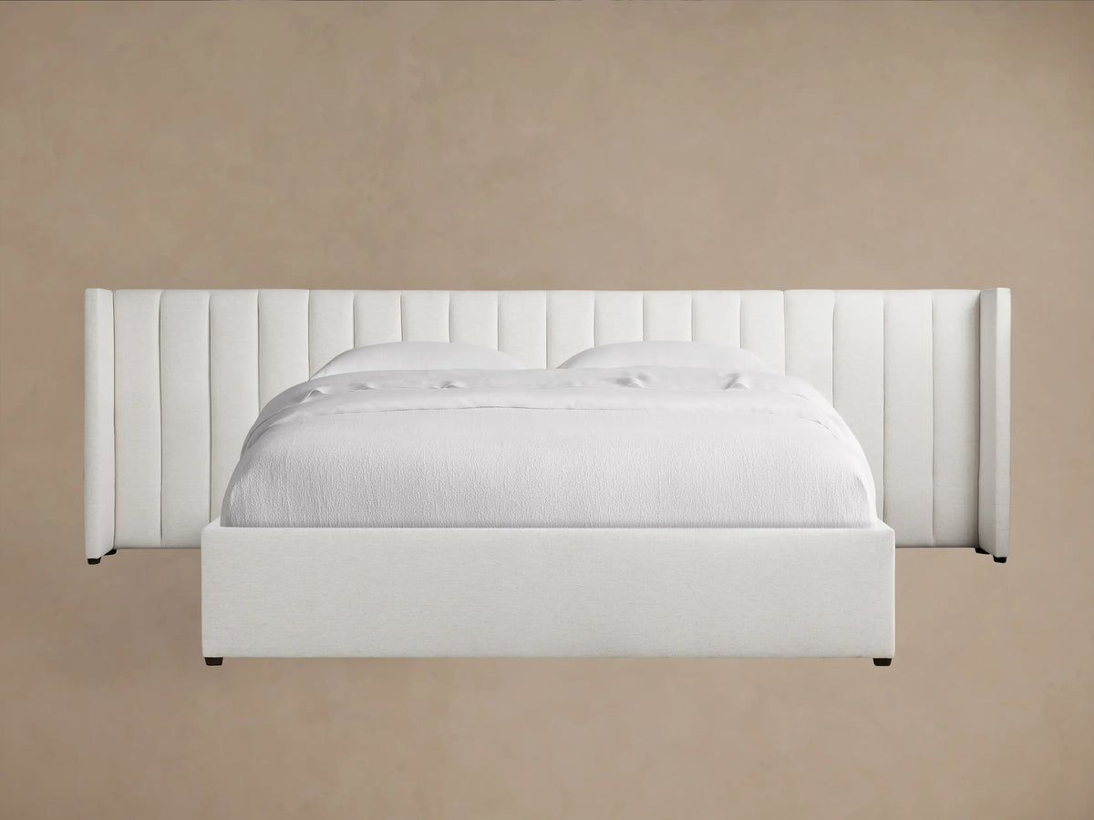 Wyller Channel-Stitched Extended Headboard Bed in white, with a low footboard, white bedding, against a tan wall & wood floor.