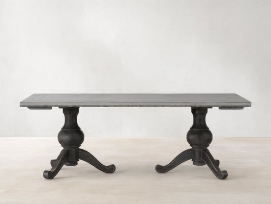 Girardi Geo Link Bell'Arte Dining Table featuring a long rectangular top in Antique White with an Antique Grey geometric border, supported by two ornate dark pedestal legs on a wooden floor.