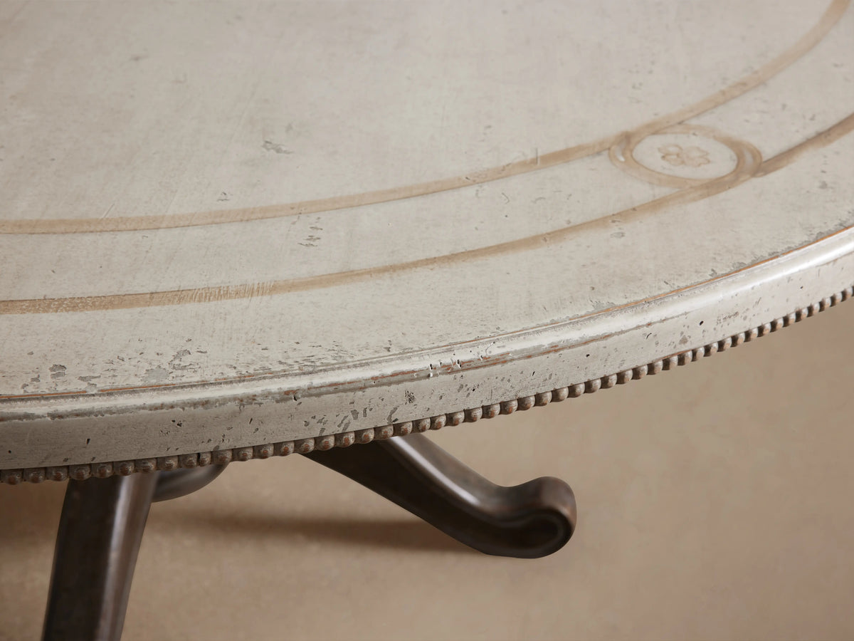 Girardi Geo Link Bell'Arte Round Dining Table in antique white with a dark, ornate pedestal base on a wooden floor.