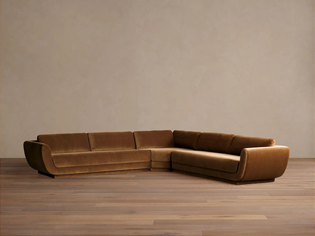 Fontana Corner Sectional, L-shaped, upholstered in warm brown velvet with curved arms, shown on a wooden floor.