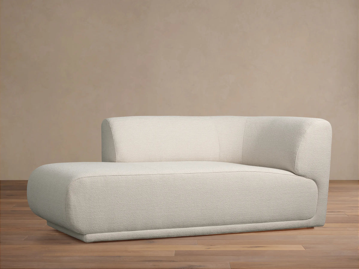Light cream Gianni Chaise with a low, rounded design and textured fabric, backrest on the right, on a wood floor.