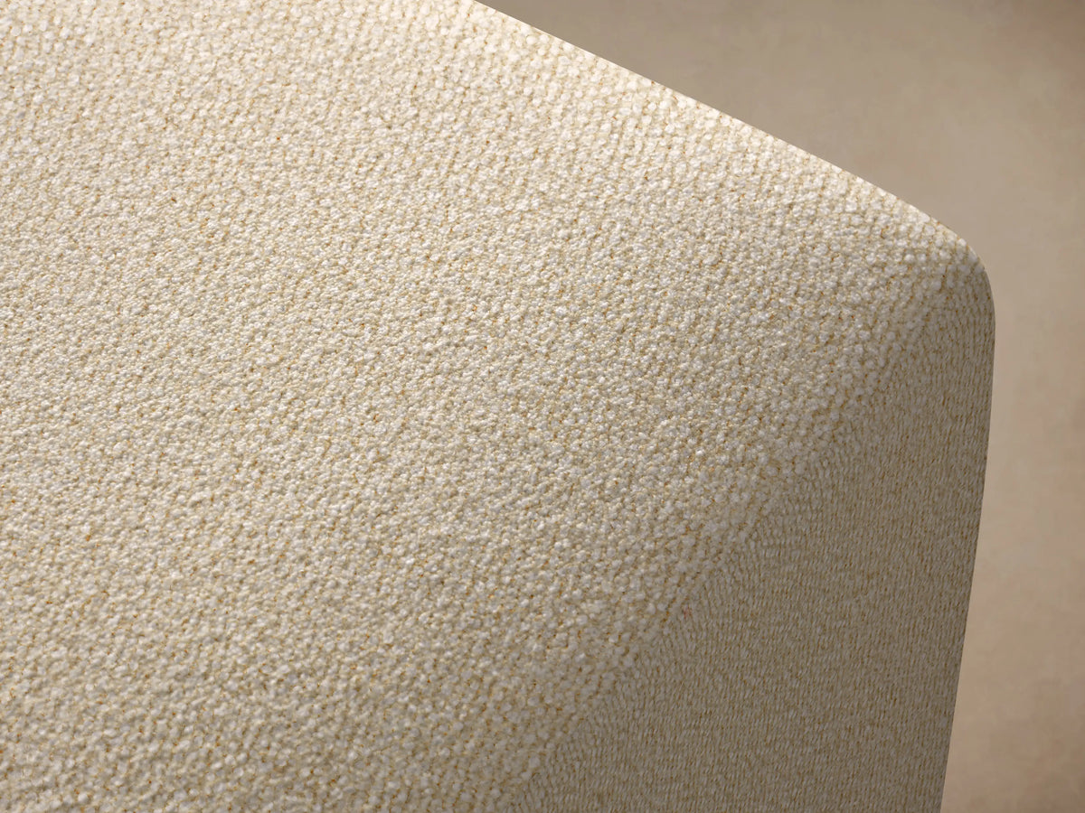 Lovina Ottoman, upholstered in light beige textured fabric, with rounded top edges, on a wood floor against a warm beige wall.