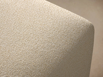Lovina Ottoman, upholstered in light beige textured fabric, with rounded top edges, on a wood floor against a warm beige wall.