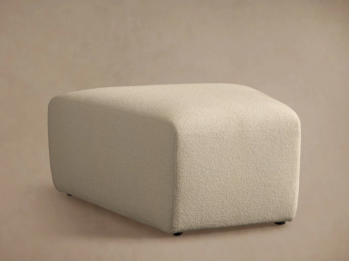 Lovina Ottoman close-up: light beige, finely textured fabric on a rounded upholstered corner, against a soft background.