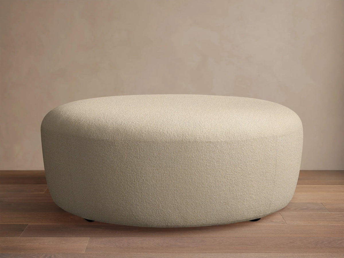 Lovina Ottoman: a rounded, light beige upholstered ottoman with textured fabric, on a wooden floor against a neutral wall.