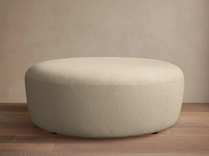 Lovina Ottoman: a rounded, light beige upholstered ottoman with textured fabric, on a wooden floor against a neutral wall.