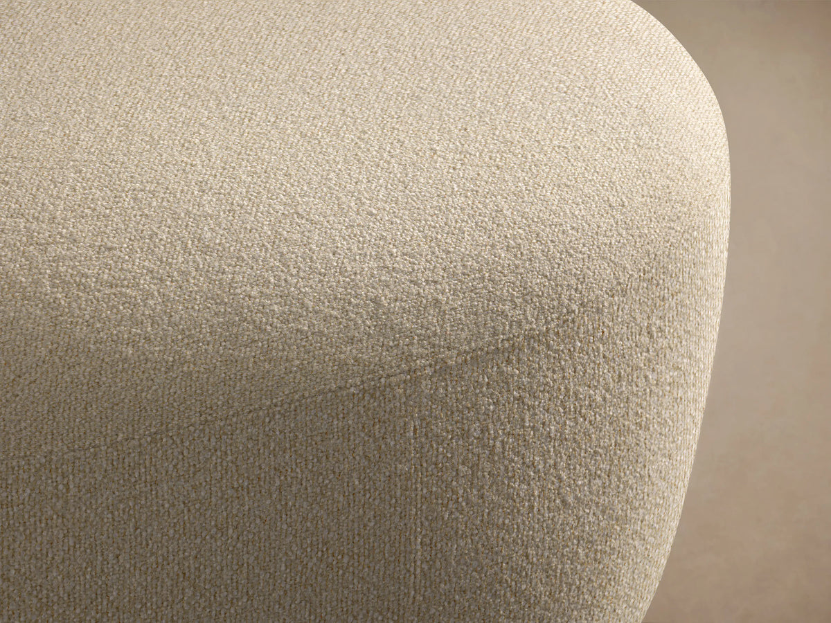 Lovina Ottoman: a rounded, light beige upholstered ottoman with textured fabric, on a wooden floor against a neutral wall.