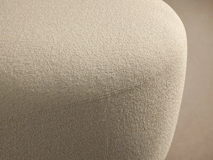 Lovina Ottoman: a rounded, light beige upholstered ottoman with textured fabric, on a wooden floor against a neutral wall.