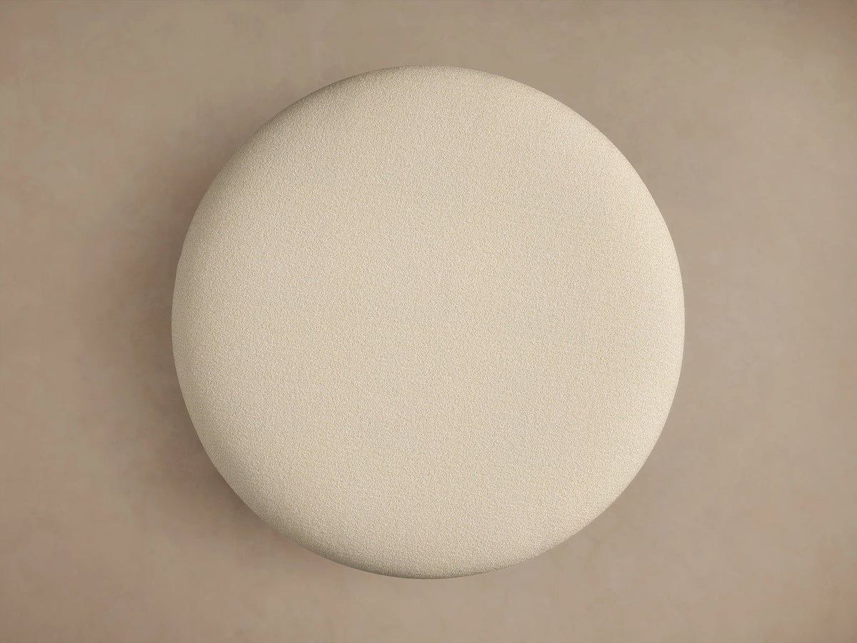 Close-up of Lovina Ottoman showing light beige, textured upholstery fabric with a subtle seam on its plush, curved surface.