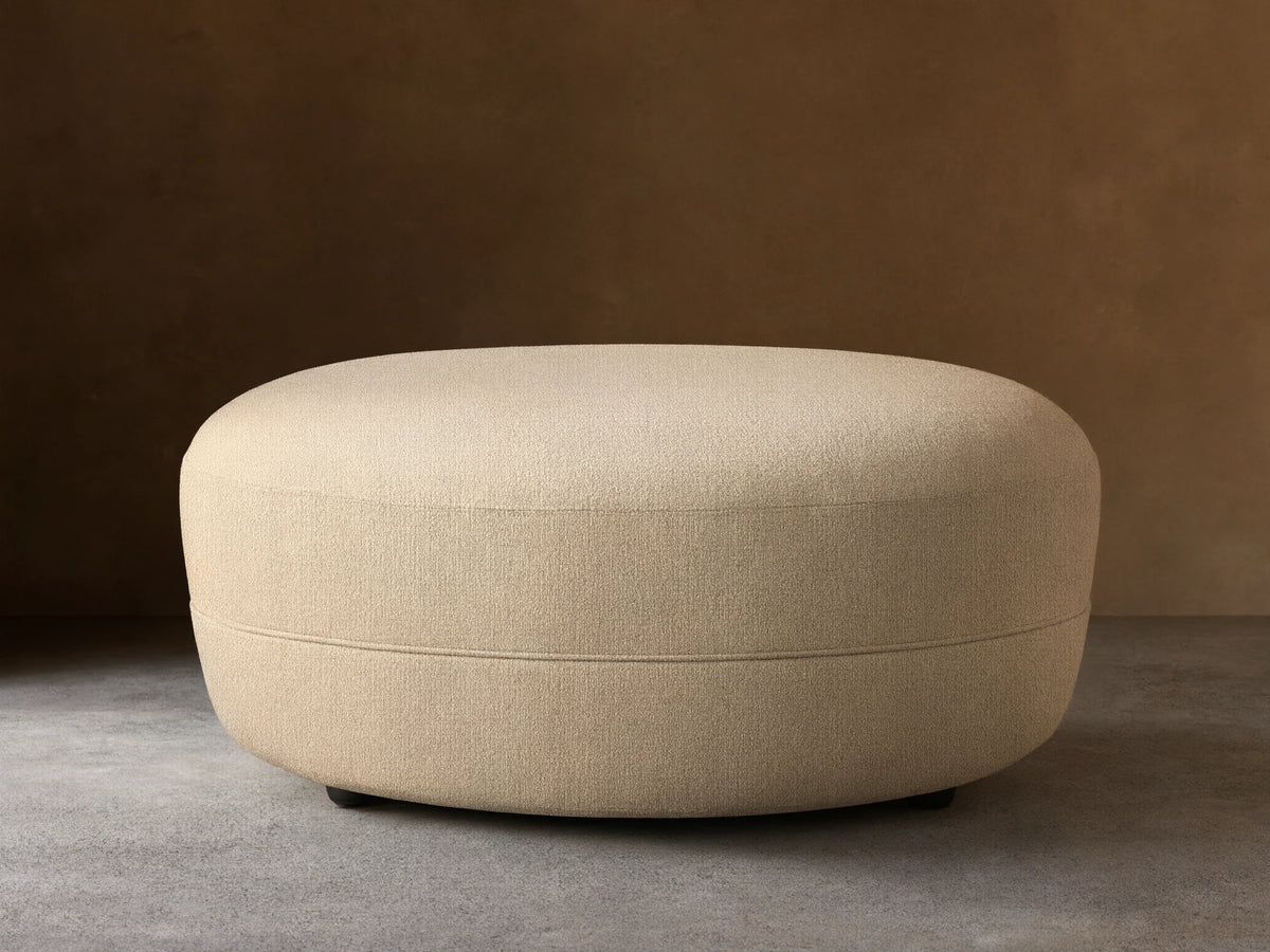 Lovina Ottoman: a rounded, light beige upholstered ottoman with textured fabric, on a wooden floor against a neutral wall.