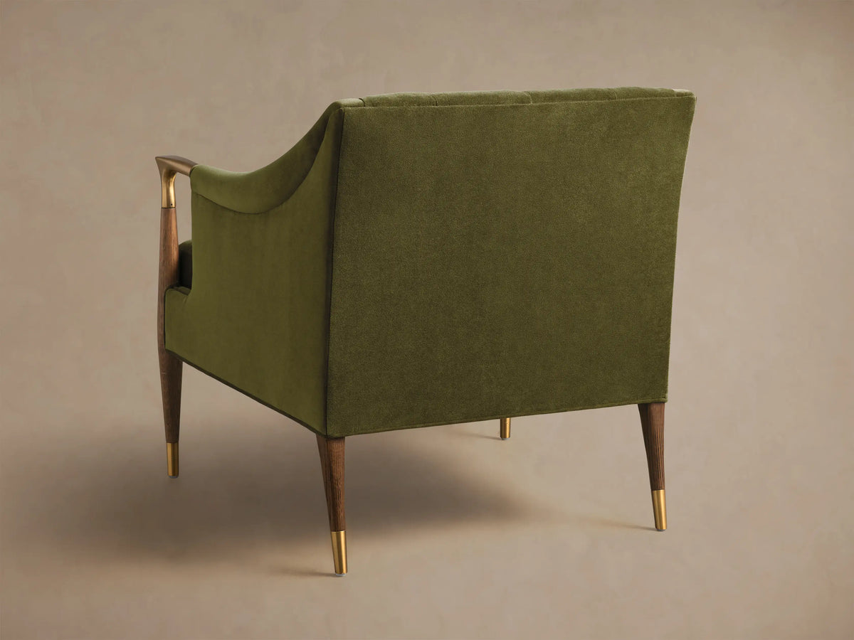 Angled front view of an olive green Fisher Club Chair with velvet upholstery, tufted back, wooden arms with brass accents, and a lumbar pillow.