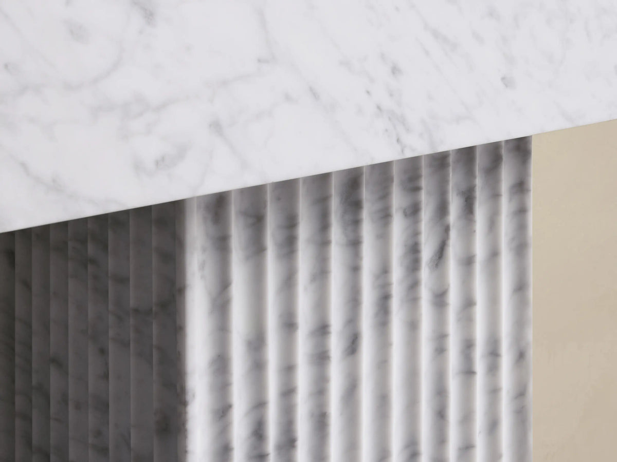 Close-up of a white marble slab with gray veins angled over a vertically fluted white marble panel, showing shadows.