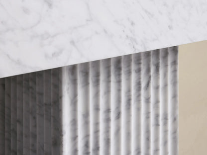 Close-up of a white marble slab with gray veins angled over a vertically fluted white marble panel, showing shadows.